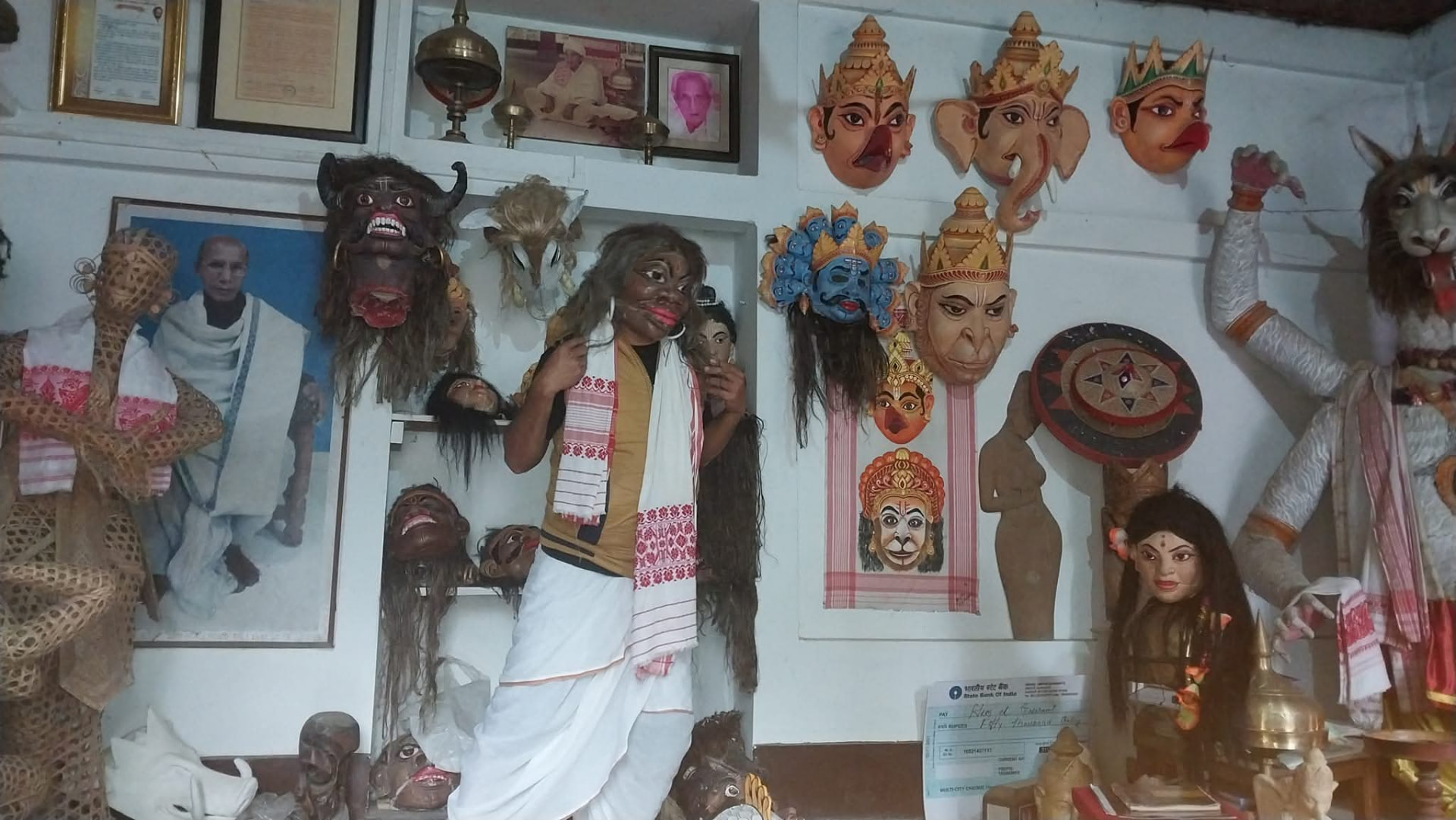 Traditional Assamese bhaona masks of deities and mythological characters displayed on the walls of a mask-making workshop, with a performer holding a mask