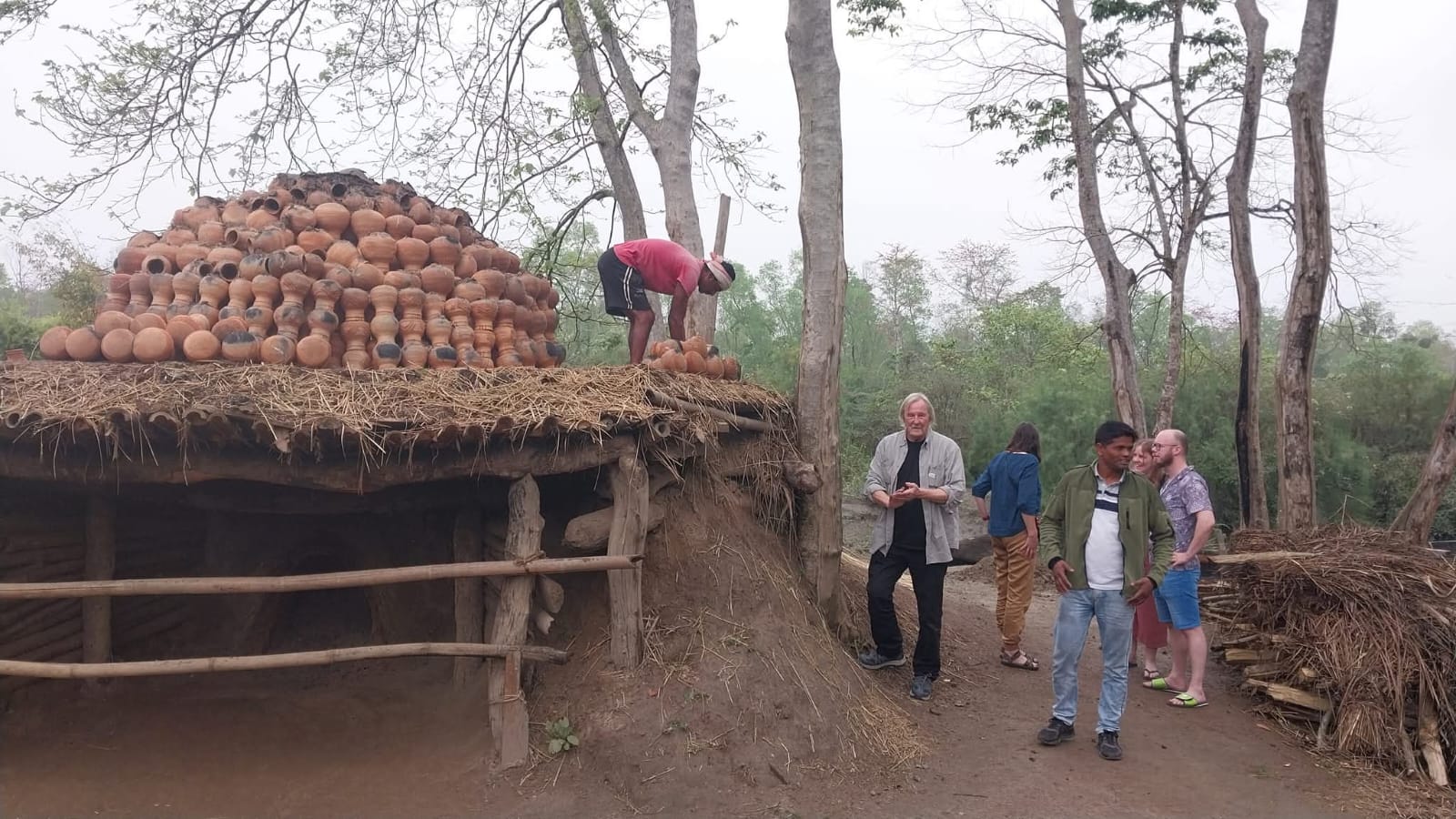 Stacked earthen pots being fired over a straw-covered open kiln in a Majuli village, with visitors watching from the path