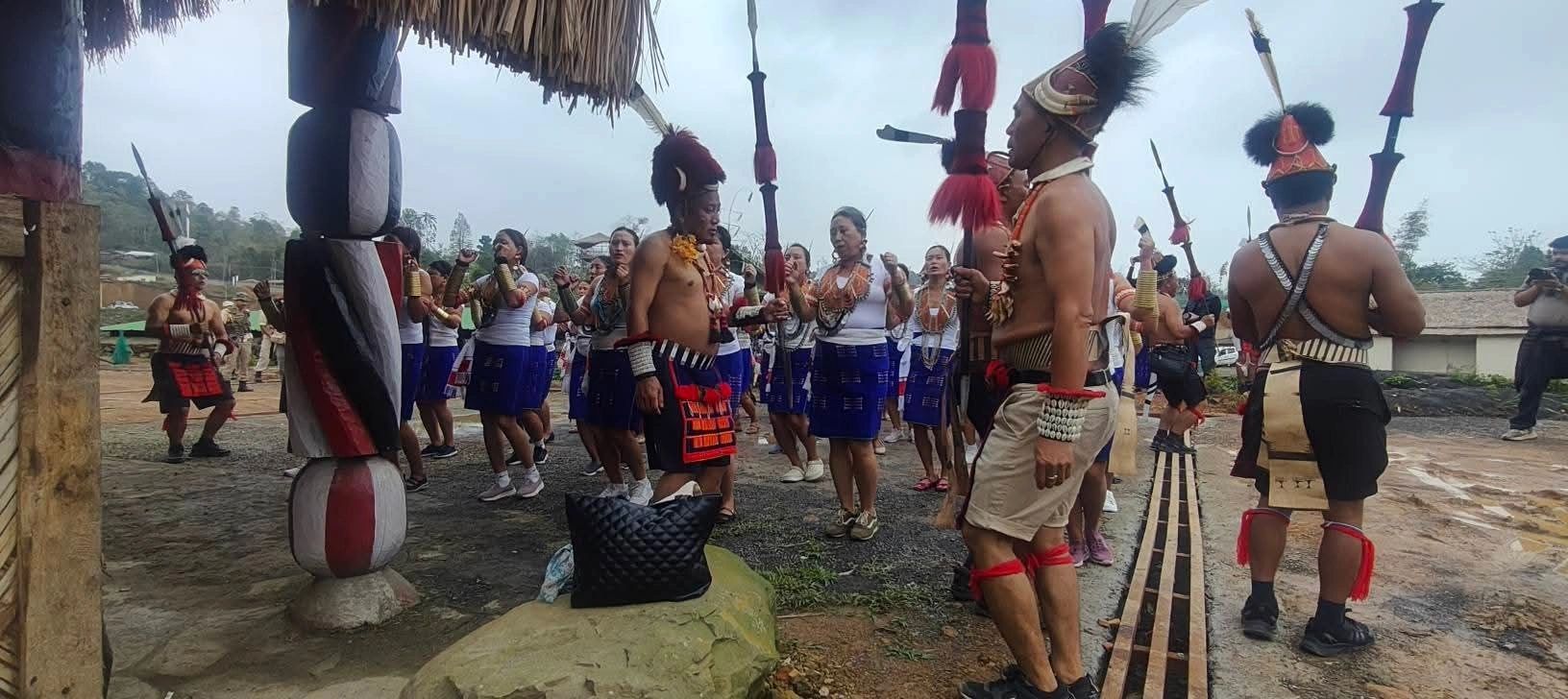 Konyak warriors in red goat-hair and feather headgear holding spears and daos beside a carved village post during Aoleang Festival