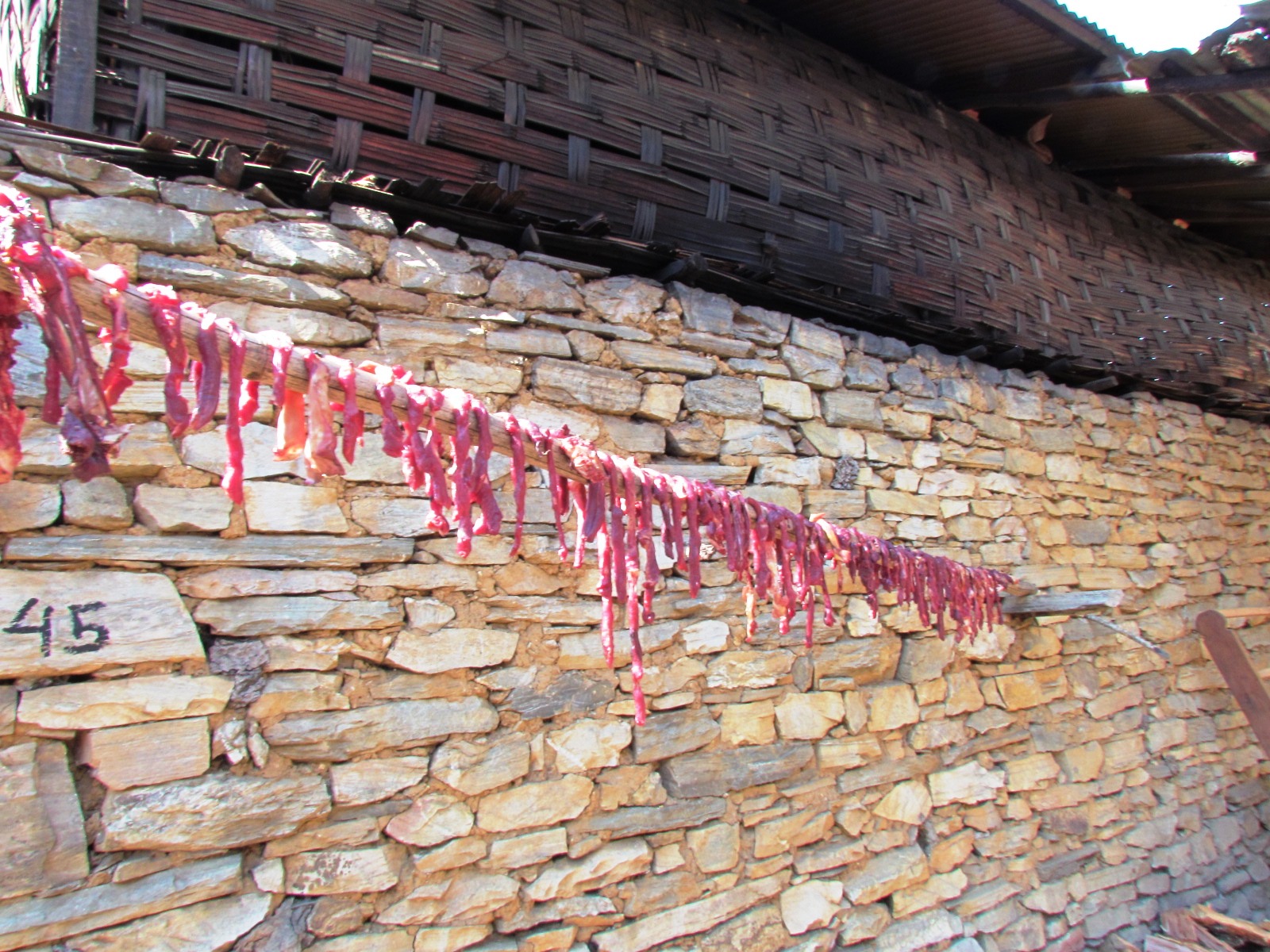 Strips of yak meat drying on a bamboo pole along a dry-stone wall below a woven bamboo gable in a Monpa village