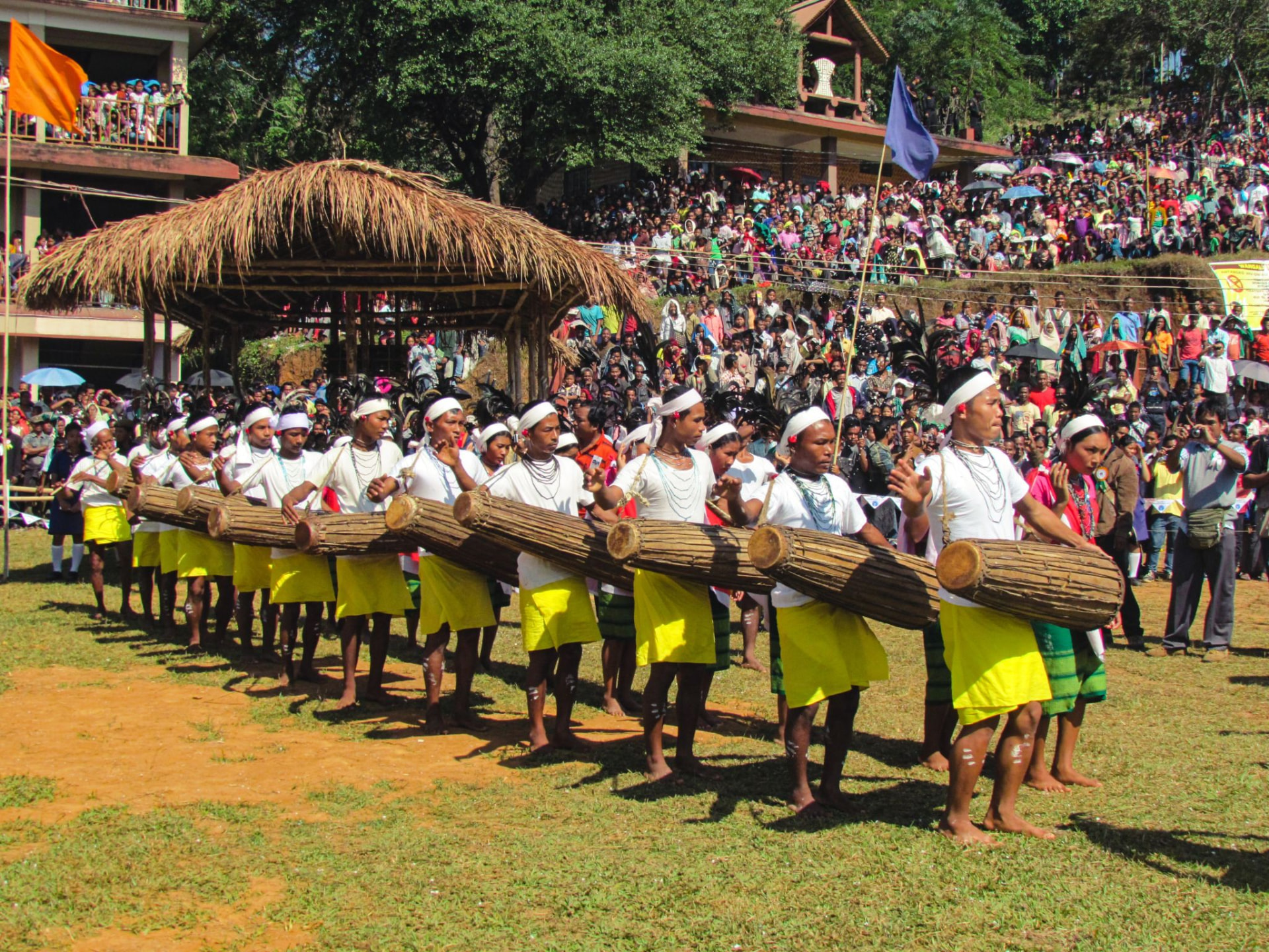 Line of drummers carrying long wooden drums at a hillside festival ground in Northeast India, watched by a large crowd