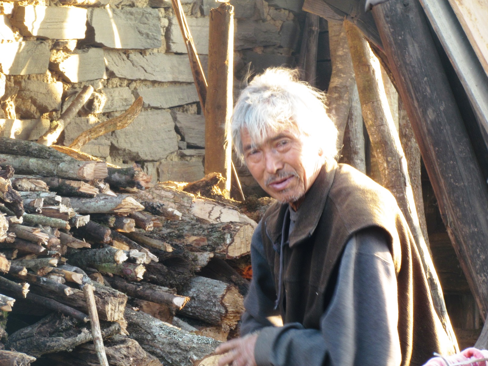 Elderly Monpa man with white hair sitting beside a stack of split firewood outside a stone house in winter