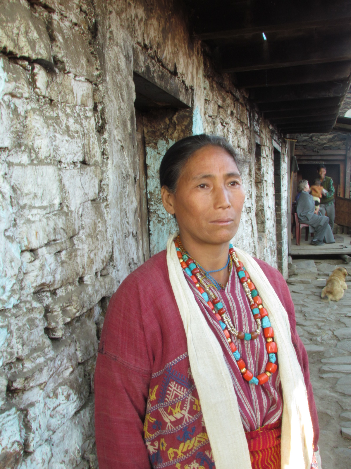 Monpa woman in a hand-loomed striped jacket wearing coral and turquoise necklaces, standing outside a whitewashed stone house