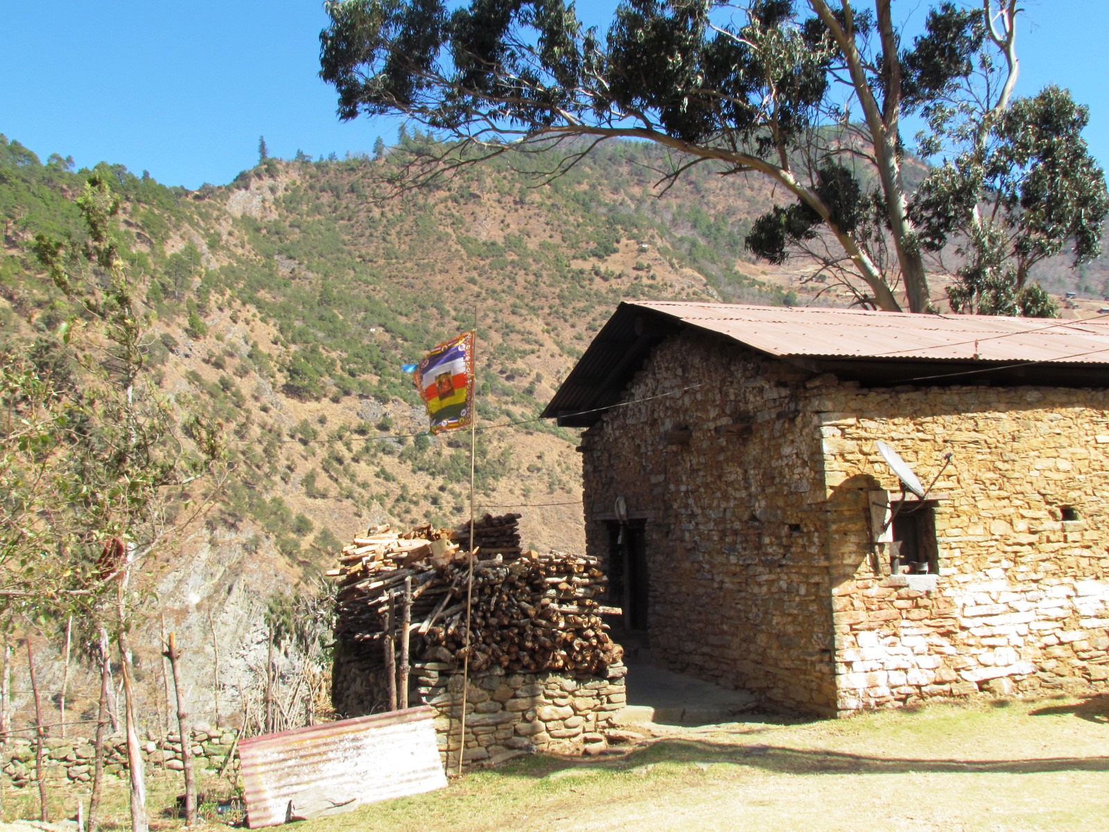 Stone Monpa farmhouse with tin roof, stacked firewood and a Buddhist prayer flag above the Sangti Valley hillside