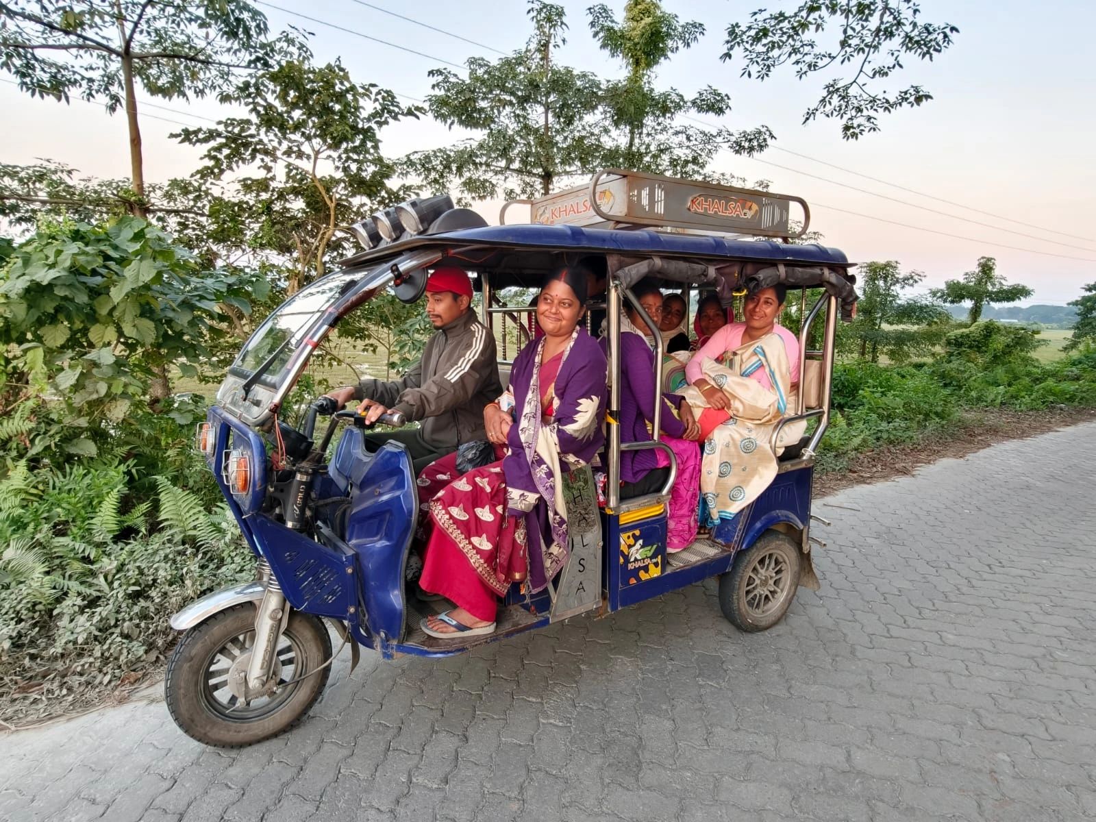 Passengers riding a blue battery-powered e-rickshaw along a rural road bordered by fields in Assam