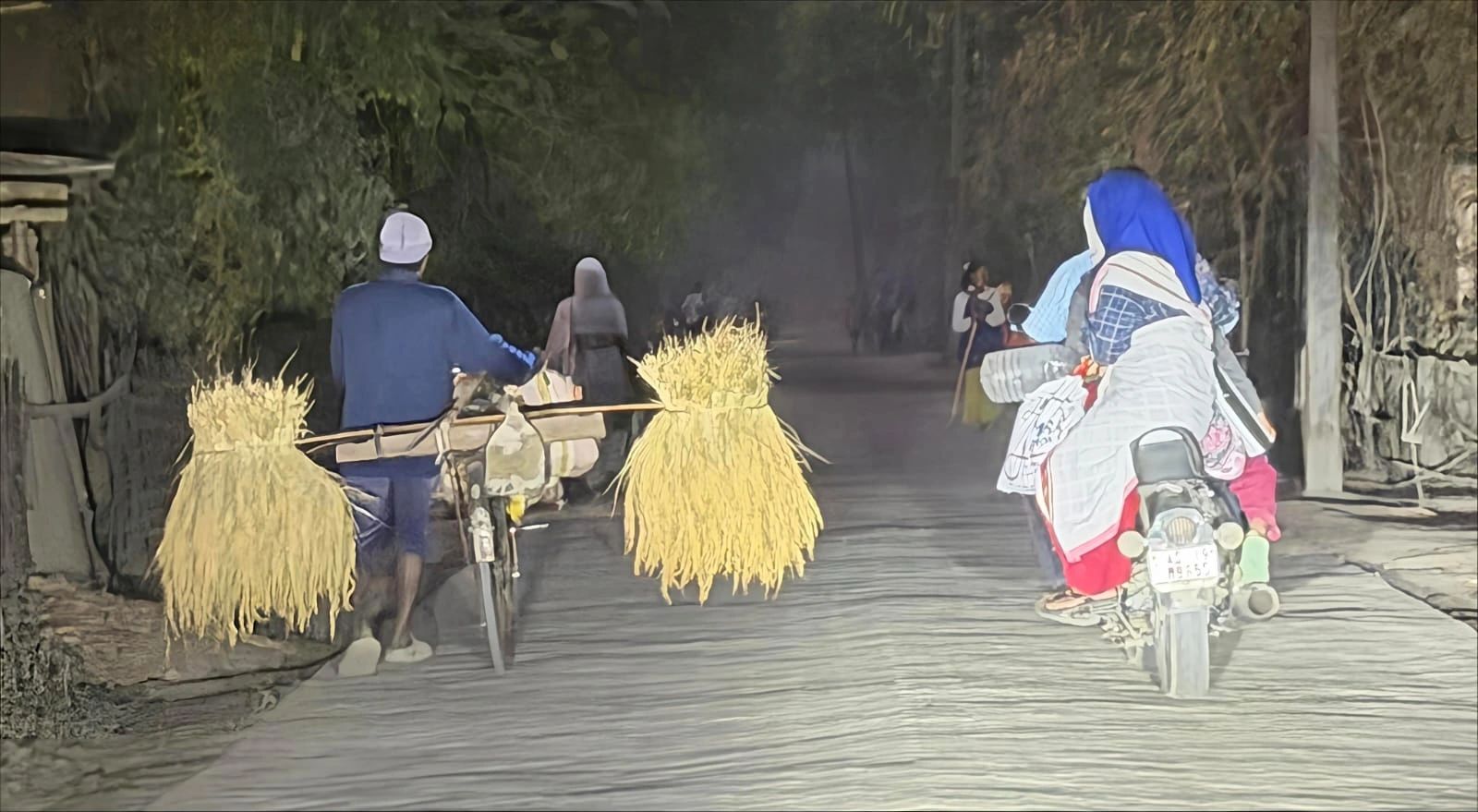 Villagers cycling and riding home along a dusty island road at dusk, carrying bundles of harvested paddy