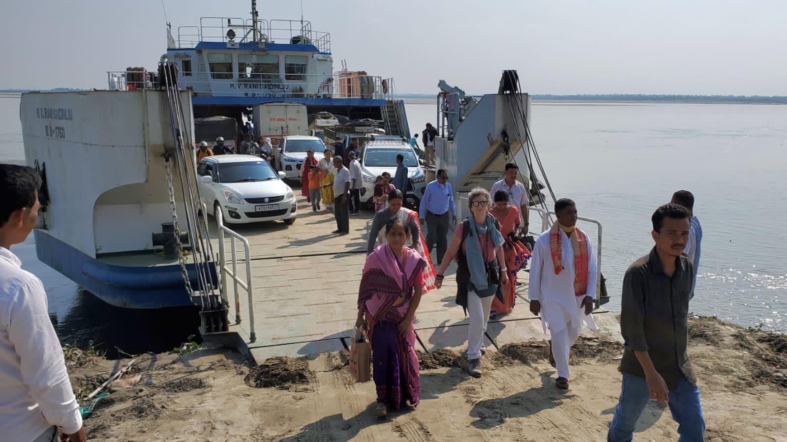 Passengers walking down the ramp of a Brahmaputra river ferry onto a sandy bank, with cars waiting to disembark