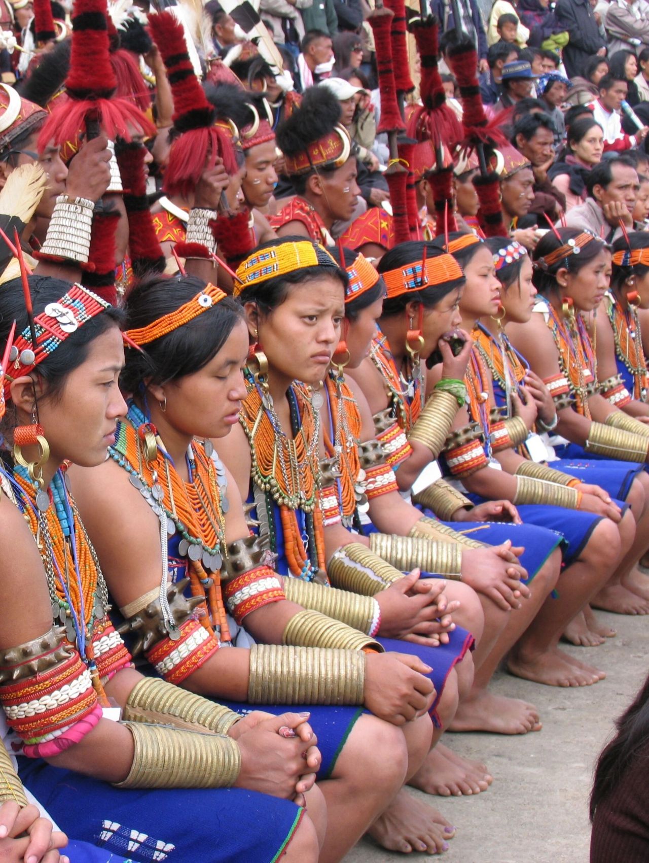 Women and men in traditional festival dress with beadwork, brass ornaments and headgear seated at a festival ground in Northeast India